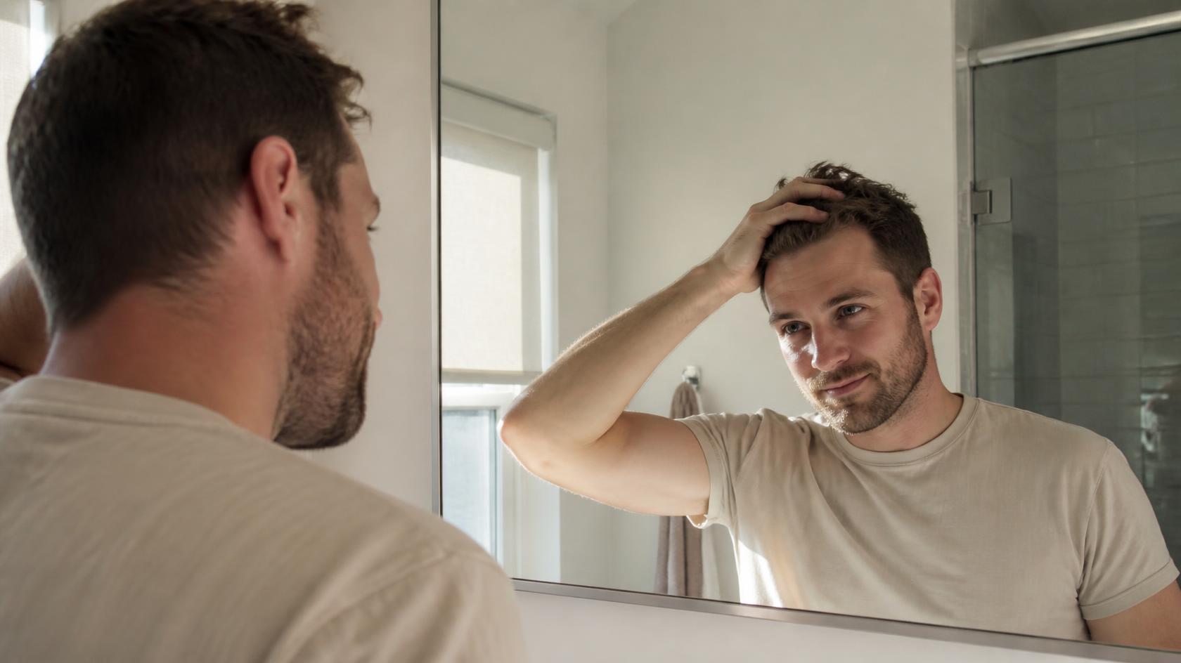 Man smiles softly at his reflection, touching his hair with quiet, hopeful confidence.